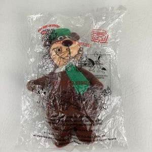 Pode incluir: Um urso de pelúcia castanho com um chapéu e cachecol verdes, selado num saco de plástico transparente. A embalagem apresenta o texto "GET ONE IN EVERY KID'S MEAL!", "CLASSIC TOONS" e "COLLECTOR SERIES I" com várias ilustrações de desenhos animados.