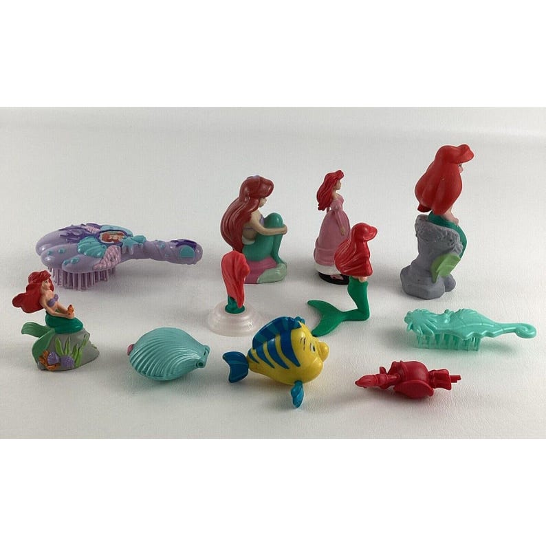 May include: A collection of colorful figurines featuring characters and items from a popular animated film. The set includes mermaid figures, a seashell, a fish, a crab, and a hairbrush. The figurines are in various poses and colors.