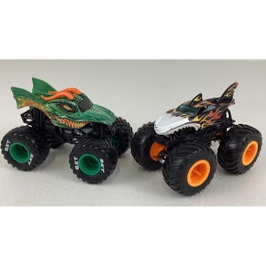 Puede incluir: Dos monster trucks de juguete. Uno es verde con un dise&ntilde;o de drag&oacute;n naranja, ruedas negras y detalles verdes. El otro es negro con un dise&ntilde;o de tibur&oacute;n, llamas naranjas y amarillas y detalles naranjas en las ruedas.