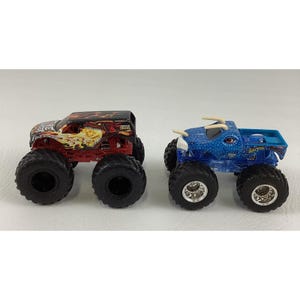 May include: Two toy monster trucks. One is red and black with a flame design, the other is blue with a textured pattern. Both have large black tires and are designed for play.