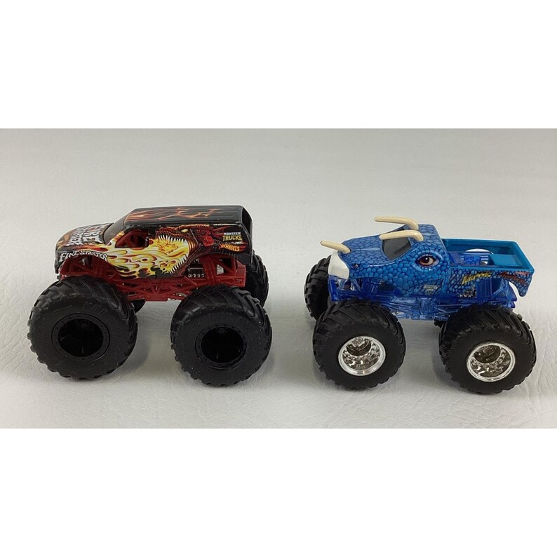 May include: Two toy monster trucks. One is red and black with a flame design, the other is blue with a textured pattern. Both have large black tires and are designed for play.