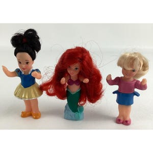 May include: Three small plastic dolls. One doll has black hair and wears a blue top and yellow skirt. Another doll has red hair and a green tail. The third doll has blonde hair and wears a pink top and blue skirt.