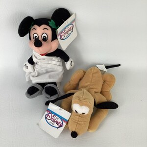 May include: A plush Mickey Mouse doll wearing a white toga and green laurel wreath, alongside a tan plush Pluto dog. Both toys feature The Disney Store tags with the logo text clearly visible. The Mickey doll has black ears and grey sandals.