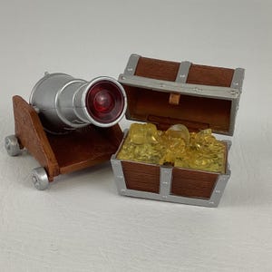 May include: A toy cannon with a silver barrel and a red lens, mounted on a brown wooden carriage with silver wheels. Next to it is an open treasure chest filled with gold-coloured plastic gems, with brown wooden sides and silver metal accents.