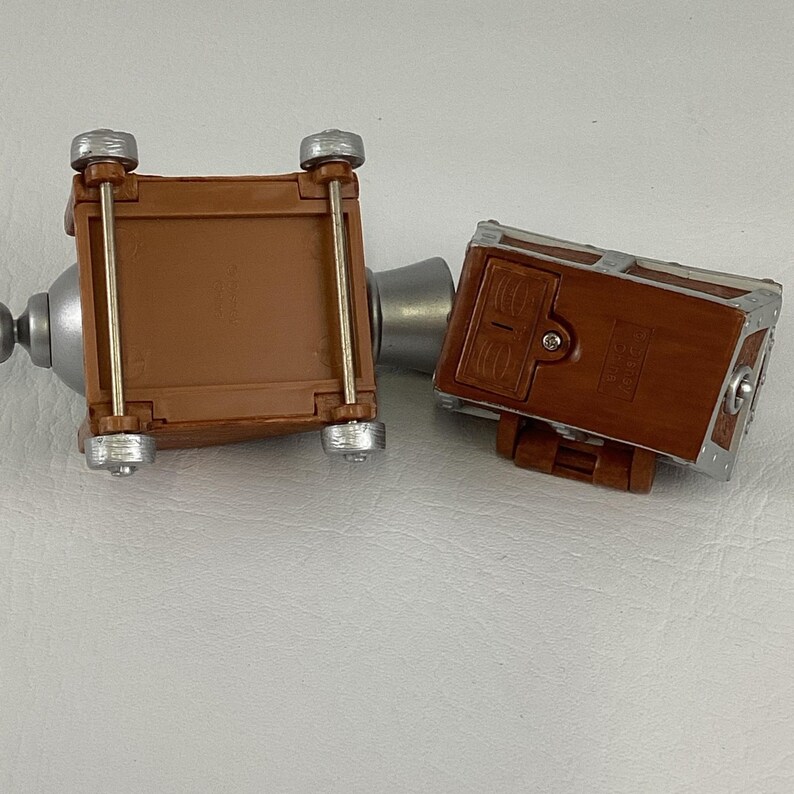 May include: Two brown toy chests with silver detailing. One chest is on its side, showing the base with four wheels. The other chest is upright, displaying the front with a latch. The chests are joined by a silver tube.