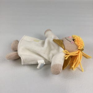 Puede incluir: Una mu&ntilde;eca de &aacute;ngel de peluche con una t&uacute;nica color crema, brazos y piernas color canela y una cara sonriente con mejillas rosadas. La mu&ntilde;eca tiene alas doradas y cabello de hilo amarillo. Una peque&ntilde;a etiqueta est&aacute; adherida a la t&uacute;nica.