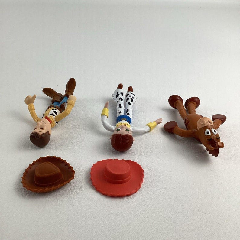 May include: Toy Story character figurines, including Woody, Jessie, and Bullseye, are displayed with their hats. Woody is in a yellow shirt, Jessie in a white shirt and cow-print pants, and Bullseye is brown. The hats are brown and red.