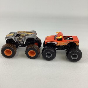 Puede incluir: Dos camiones monstruo de juguete. Uno es plateado y negro con ruedas naranjas. El otro es naranja con un dise&ntilde;o de toro y el texto "El Toro Loco".