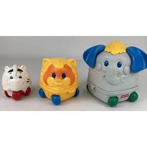 Puede incluir: Tres juguetes de animales Fisher-Price coloridos. Un tigre blanco con ruedas rojas, un león amarillo con ruedas azules y un elefante gris con orejas azules y ruedas verdes. El elefante tiene una corona amarilla.
