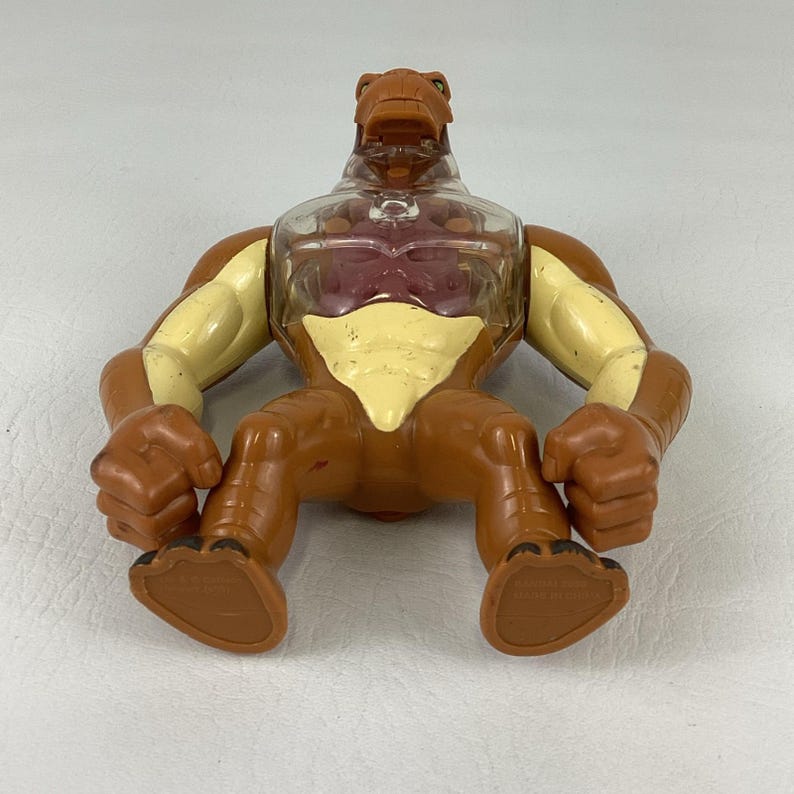 May include: A brown and cream-colored action figure featuring a clear plastic chest plate that reveals a dark red interior structure. The figure has a muscular build with large arms, clenched fists, and a reptilian-like head with an open mouth.