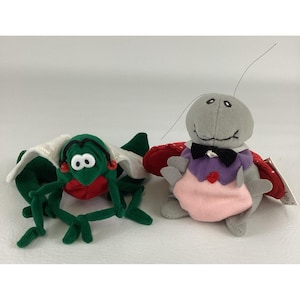 Peut inclure: Deux jouets en peluche : une créature verte et rouge en forme de bug avec de grands yeux et une cape blanche, et un insecte gris avec un nœud papillon et une tenue rose et violette. Les jouets sont sur une surface blanche.