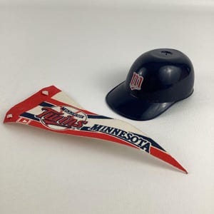 Op de afbeelding: Een blauw-witte Minnesota Twins-vlag met een honkbal en het logo van het team. Een blauwe plastic honkbalpet met een witte 'M' op de voorkant.