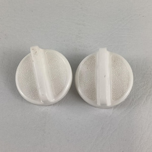 May include: Two white, circular appliance knobs with a textured surface and a raised, vertical bar. The knobs have a small, raised tab at the top. The knobs are set against a light gray background.