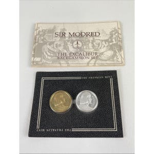Puede incluir: Un juego de backgammon con dos fichas redondas, una dorada y otra plateada, presentadas sobre una tarjeta negra. La tarjeta lleva la inscripción "THE FRANKLIN MINT". Encima, un papel doblado dice "SIR MODRED" y "THE EXCALIBUR BACKGAMMON SET".