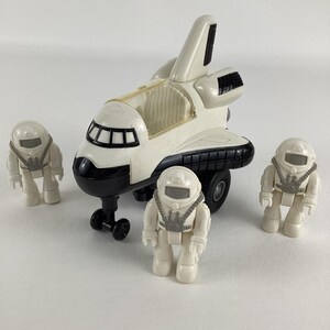 May include: A white and black toy space shuttle with a clear cockpit and four matching astronaut figures. The shuttle has black wheels and a black nose. The astronauts are wearing white space suits with gray accents.