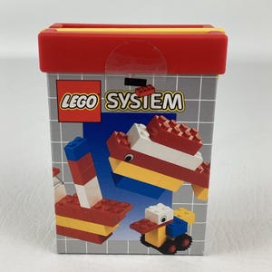 May include: A rectangular LEGO System storage box with a red lid and a gray body. The box features the LEGO logo and illustrations of assembled LEGO creations in red, yellow, white, and blue. The box is designed to store LEGO bricks.