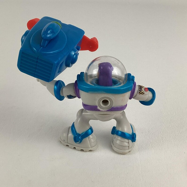 Op de afbeelding: Plastic speelgoedfiguur van een ruimteranger in wit, blauw en paars. De figuur houdt een blauw en rood apparaat vast. Het speelgoed heeft een doorzichtige helm en zit in een hurkende positie. Kinderspeelgoed.