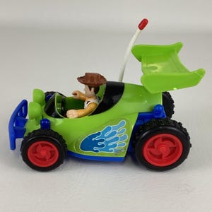 May include: A green and blue toy race car with red wheels and a spoiler. The car features a Woody figure in the driver's seat and a remote control antenna. The car has a blue and white design on the side.