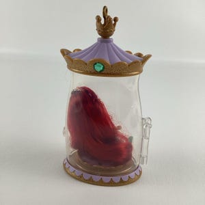 Puede incluir: Un recipiente de pl&aacute;stico transparente con tapa y base decorativas, que contiene una mu&ntilde;eca pelirroja en su interior. La tapa es morada con ribete dorado y una peque&ntilde;a corona en la parte superior. Una gema verde est&aacute; engastada en el centro de la tapa.