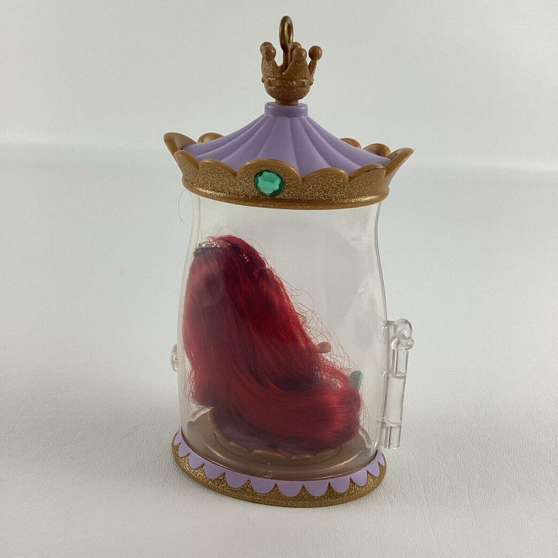 Puede incluir: Un recipiente de pl&aacute;stico transparente con tapa y base decorativas, que contiene una mu&ntilde;eca pelirroja en su interior. La tapa es morada con ribete dorado y una peque&ntilde;a corona en la parte superior. Una gema verde est&aacute; engastada en el centro de la tapa.