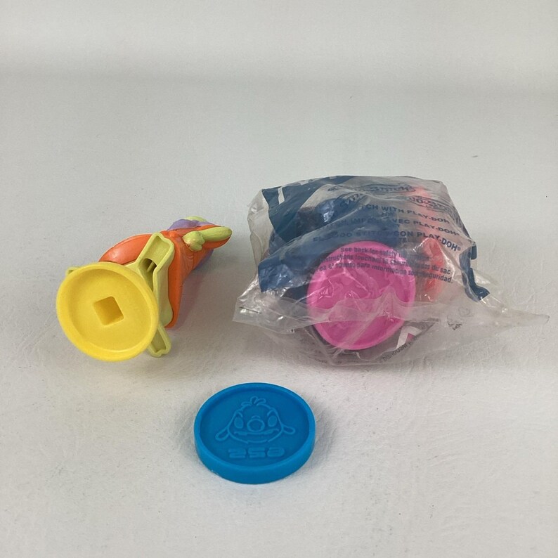 May include: A collection of colorful children's toys, including a blue disc with a cartoon character, a yellow and orange toy, and a Play-Doh container with pink and orange play dough inside a plastic bag.