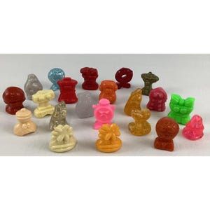 May include: A collection of small, colorful plastic figurines in various shapes. The figurines are in shades of red, orange, yellow, green, pink, blue, and gray. Some have abstract designs, while others resemble animals or objects.