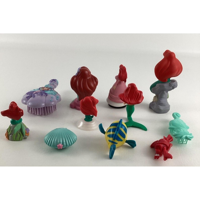 May include: A collection of colorful miniature figurines featuring characters and elements from a popular animated film. The set includes a variety of figures, such as a seashell, a turtle, and several red-haired characters, all in vibrant colors.