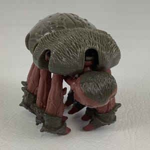 K&ouml;nnte beinhalten: Eine Kunststoff-Actionfigur einer Kreatur mit einem gro&szlig;en, abgerundeten, olivgr&uuml;nen Panzer mit gepr&auml;gten Mustern. Die Figur hat mehrere r&ouml;tlich-braune, texturierte Gliedma&szlig;en, die in dunkelolivgr&uuml;nen, gepanzerten Manschetten enden.