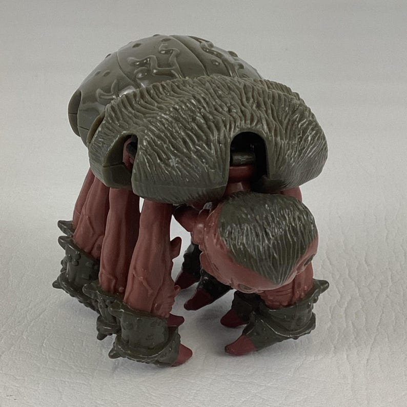 K&ouml;nnte beinhalten: Eine Kunststoff-Actionfigur einer Kreatur mit einem gro&szlig;en, abgerundeten, olivgr&uuml;nen Panzer mit gepr&auml;gten Mustern. Die Figur hat mehrere r&ouml;tlich-braune, texturierte Gliedma&szlig;en, die in dunkelolivgr&uuml;nen, gepanzerten Manschetten enden.
