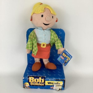 以下が含まれることがあります： 「Bob the Builder」シリーズのウェンディのぬいぐるみ。ウェンディは水色のシャツ、オレンジ色のスカート、緑のチェック柄のジャケットを着ています。人形は、Bob the Builderのロゴとウェンディの名前が入った青いディスプレイスタンドに立っています。