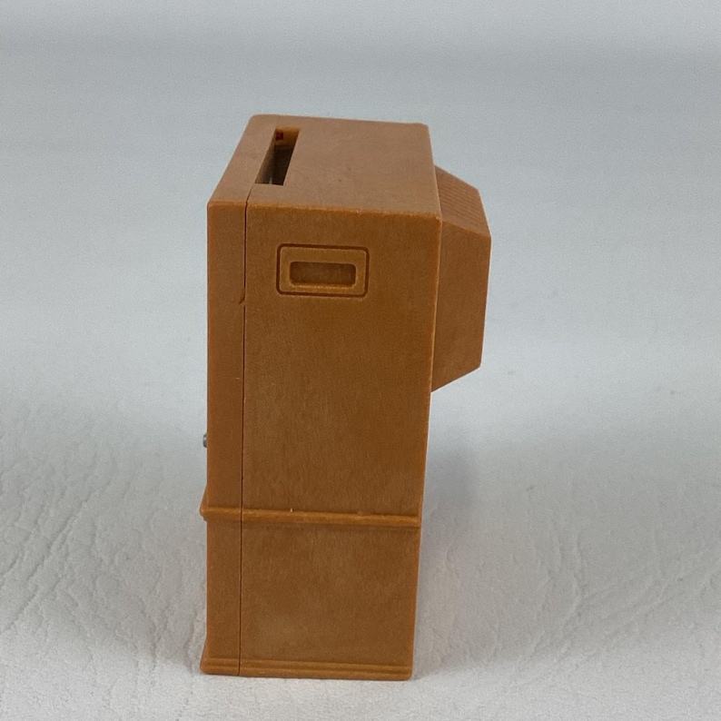 Op de afbeelding: Een kleine, bruine speelgoedtelevisie in vintage-stijl. De speelgoed-tv heeft een rechthoekige vorm met een sleuf aan de bovenkant en een rechthoekige opening aan de voorkant. Het ontwerp doet denken aan oudere televisies.
