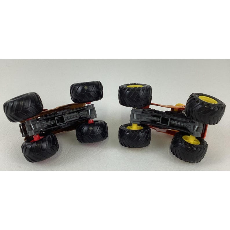 May include: Two toy monster trucks. Each truck has large black tyres with yellow rims, a red chassis, and a brown body. The trucks are positioned on a white surface, with one slightly angled towards the other.