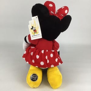 Pu&ograve; includere: Un peluche di Minnie Mouse, vista da dietro, con un vestito rosso a pois bianchi, un fiocco abbinato e scarpe gialle. Il giocattolo ha una testa e orecchie nere. Un'etichetta con Topolino e "Walt Disney World" &egrave; attaccata.
