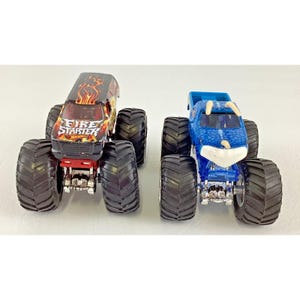 May include: Two toy monster trucks. One is black with red and orange flames and the text "Fire Starter". The other is blue with a white front. Both have large black tires.