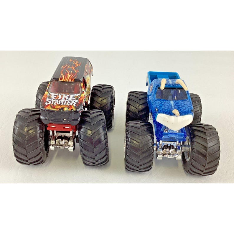 May include: Two toy monster trucks. One is black with red and orange flames and the text "Fire Starter". The other is blue with a white front. Both have large black tires.