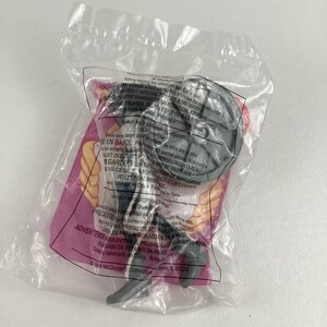 K&ouml;nnte beinhalten: Ein versiegelter Plastikbeutel mit einer grauen Plastikspielfigur. Die Figur hat einen kreisf&ouml;rmigen Kopf und einen langen, d&uuml;nnen K&ouml;rper. Die Verpackung enth&auml;lt Text in mehreren Sprachen, darunter Englisch und Spanisch.