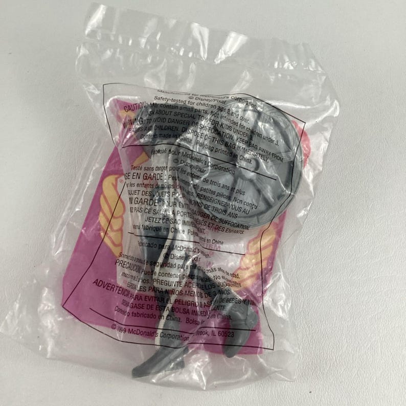 K&ouml;nnte beinhalten: Ein versiegelter Plastikbeutel mit einer grauen Plastikspielfigur. Die Figur hat einen kreisf&ouml;rmigen Kopf und einen langen, d&uuml;nnen K&ouml;rper. Die Verpackung enth&auml;lt Text in mehreren Sprachen, darunter Englisch und Spanisch.
