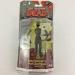 May include: A McFarlane Toys action figure of Michonne's Pet Zombie Mike from The Walking Dead, sealed in original blister packaging. The figure features a gray, decaying torso with a metal chain around the neck and dark blue pants.