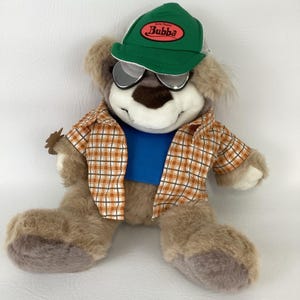 Puede incluir: Un oso de peluche que lleva una gorra verde con la palabra "Bubba", gafas de sol, una camisa a cuadros y una camiseta azul. El oso es de color marrón claro con una nariz y boca marrón oscuro. El oso está sentado.