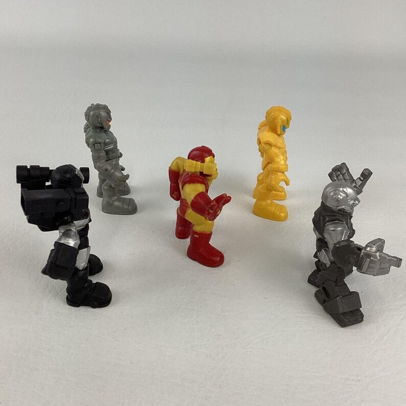 May include: A collection of five small action figures in various colors. The figures include red and yellow, silver, black, and yellow. The figures are standing on a white surface.