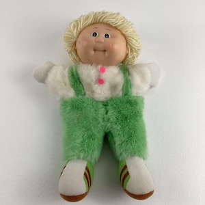 Puede incluir: Una muñeca de peluche Cabbage Patch Kid con cabello rubio, vistiendo una camisa blanca con un mono verde y zapatos verdes.