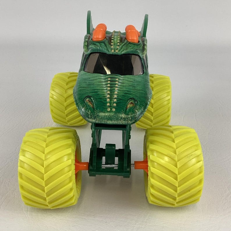 Pu&ograve; includere: Un giocattolo di monster truck verde con un design a testa di dinosauro. Il camion ha grandi pneumatici gialli e dettagli arancioni. La cabina &egrave; verde con un parabrezza nero. Il giocattolo &egrave; progettato per assomigliare a un dinosauro.