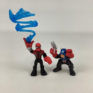 May include: Two action figures: Spider-Man in red and black with a blue energy blast, and Captain America in blue, red, and white, holding a silver gun. Both figures are in dynamic poses, ready for action.