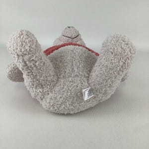 May include: A light gray plush teddy bear lying on its back with its arms and legs outstretched. The bear has a red ribbon around its neck and a small tag on its rear. The bear's fur is soft and fluffy.