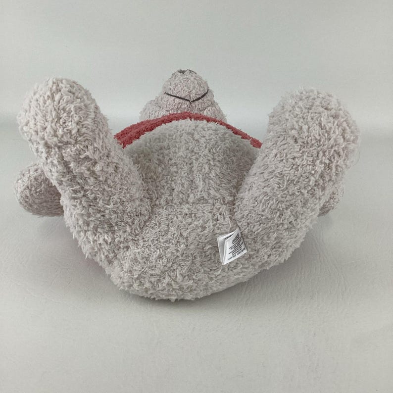 May include: A light gray plush teddy bear lying on its back with its arms and legs outstretched. The bear has a red ribbon around its neck and a small tag on its rear. The bear's fur is soft and fluffy.