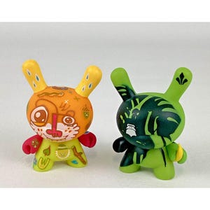 May include: Two colorful vinyl toy figures shaped like rabbits. One is orange and yellow with a variety of designs, the other is green with black leaf patterns. Both have large ears and are standing upright.