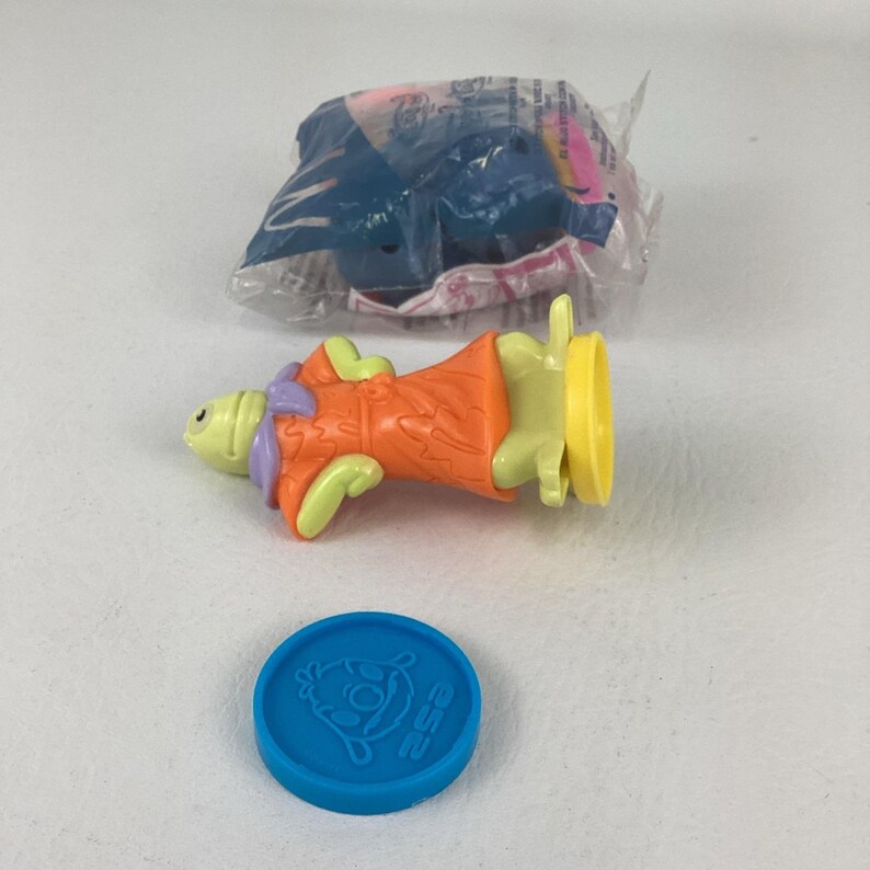 May include: A colorful plastic toy figure of a character in an orange outfit with green arms and a yellow base. A blue coin with a character illustration and the number 252 is also visible. A wrapped toy is in the background.