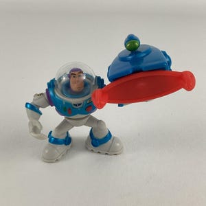 Op de afbeelding: Een Buzz Lightyear actiefiguur uit de Toy Story franchise. De figuur is voornamelijk wit, met blauwe en groene accenten. Het heeft een doorzichtige helm en houdt een rood en blauw accessoire vast.