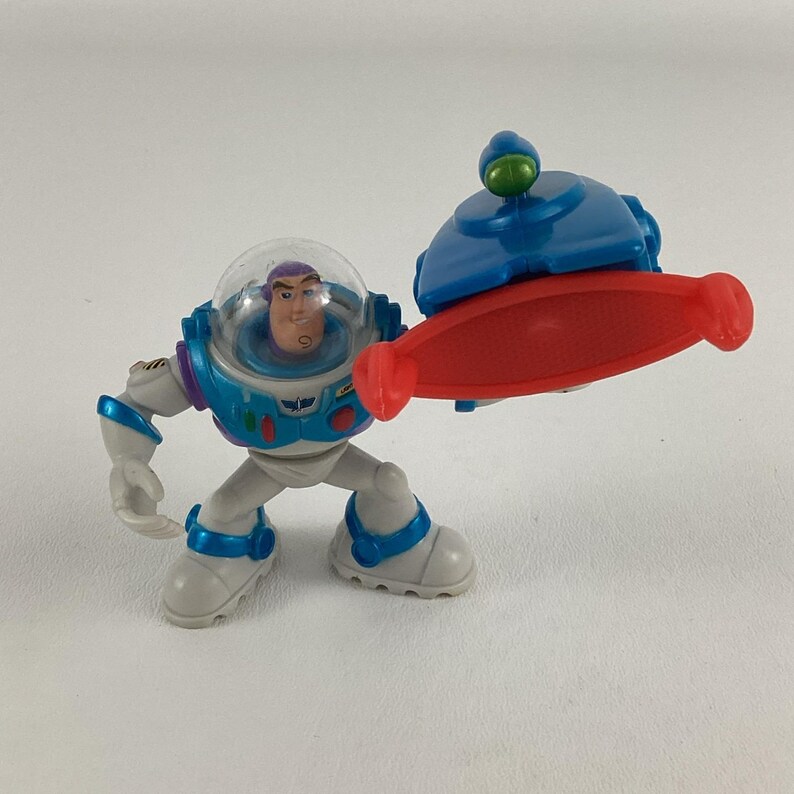 Op de afbeelding: Een Buzz Lightyear actiefiguur uit de Toy Story franchise. De figuur is voornamelijk wit, met blauwe en groene accenten. Het heeft een doorzichtige helm en houdt een rood en blauw accessoire vast.