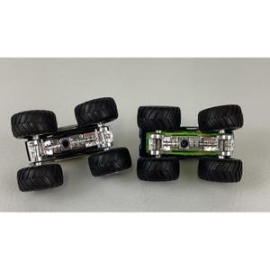 K&ouml;nnte beinhalten: Zwei Spielzeug-Monstertrucks, einer schwarz und einer blau, sind von unten zu sehen. Jeder Truck hat gro&szlig;e, schwarze, grob profilierte Reifen und ein detailliertes silbernes Fahrgestell. Die Trucks stehen auf einer hellgrauen Oberfl&auml;che.
